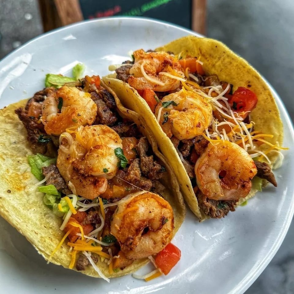 Two soft tacos filled with grilled shrimp, sliced steak, lettuce, tomatoes, avocado, and shredded cheese on a plate.